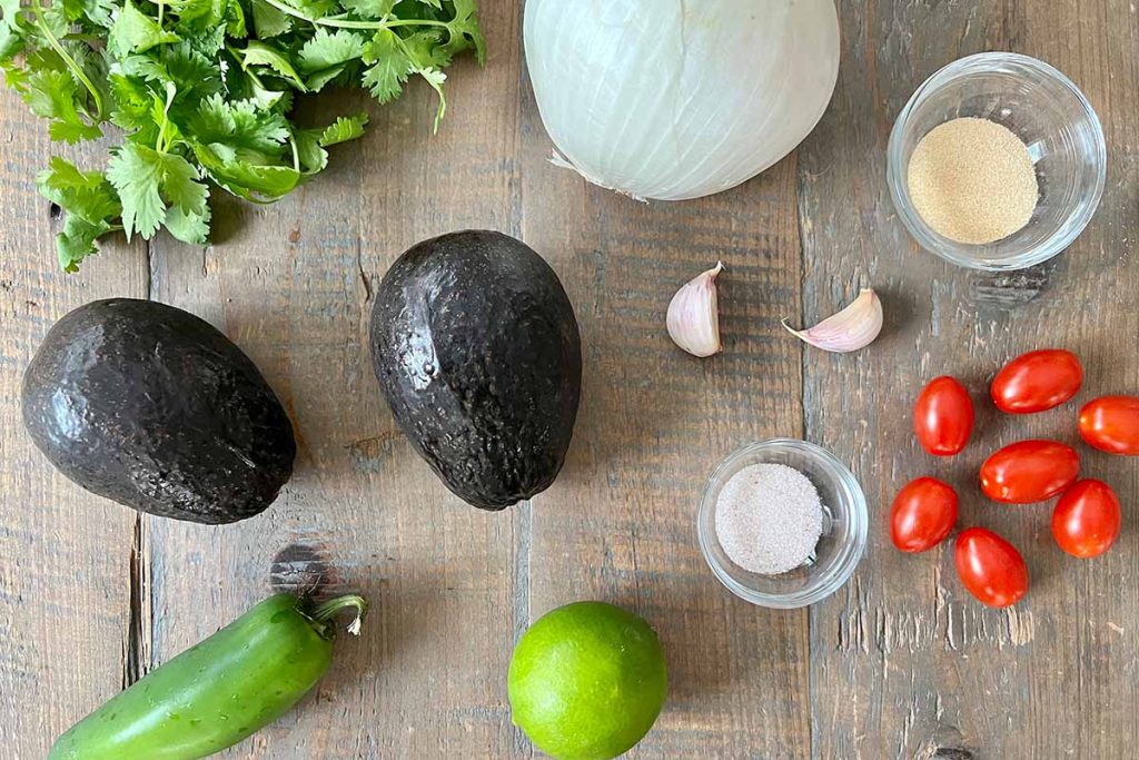 fresh whole guacamole ingredients (avocado, tomato, onion, cilantro, garlic, sea salt, lime, and jalapeno) on an antique farmhouse table made of reclaimed wood