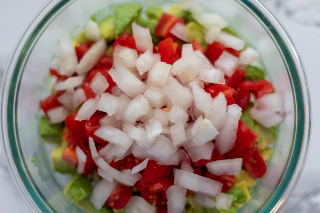 diced onion on top of tomato and avocado in a glass bowl on a marble background