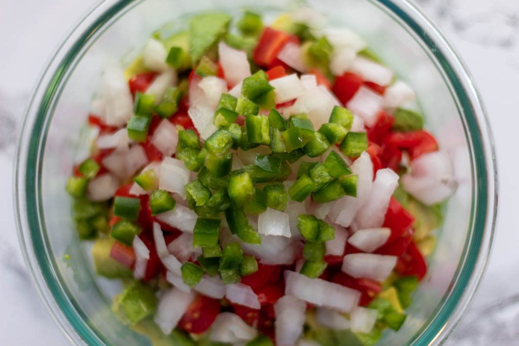 diced jalapeno on top of onion, tomato, and avocado in a glass bowl on a marble background