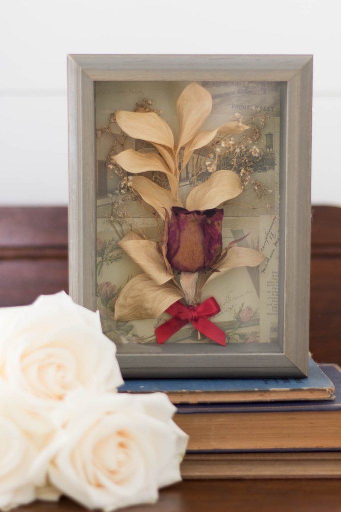 closeup of diy dried flower shadow box sitting on antique books with fresh white roses on top of an antique dresser.