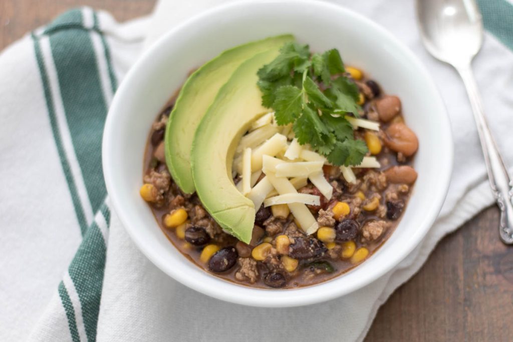 bowl filled with meat, pinto beans, black beans, corn, tomatoes, shredded cheese, cilantro, and garnished with avocado