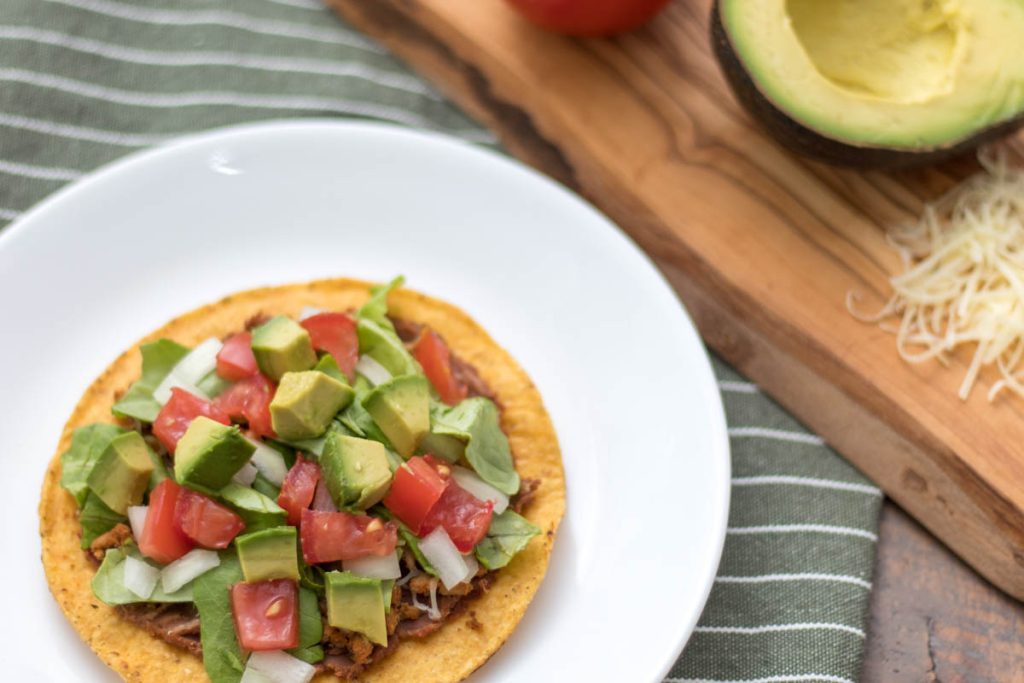 closeup of tostada shell with refried beans, taco meat, shredded cheese, lettuce, tomato, onion, and avocado on a plate next to freshly grated raw cheddar cheese, half an avocado, and whole tomato on an olive wood cutting board