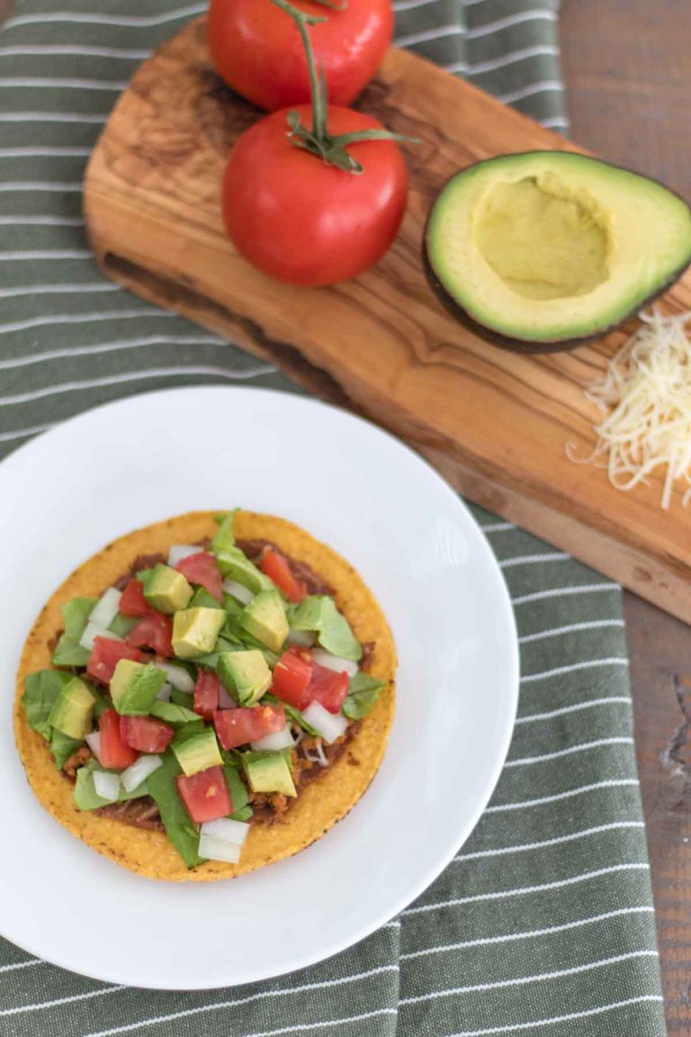 tex-mex chalupa with tostada, refried beans, taco meat, shredded cheese, lettuce, tomato, onion, and avocado on a plate next to freshly grated raw cheddar cheese, half an avocado, and whole tomato on an olive wood cutting board