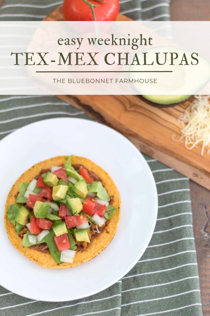 text reads"easy weeknight tex-mex chalupas." Tostada shell topped with refried beans, shredded cheese, lettuce, tomato, onion, and avocado on a white plate next to a wood cutting board and green and white striped fabric