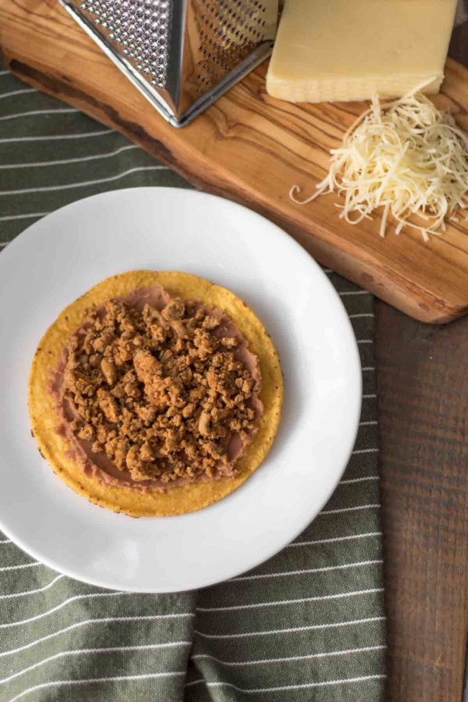 partially assembled chalupa with a tostada, refried beans and taco meat on a plate next to freshly grated raw cheddar cheese and cheese grater on an olive wood cutting board