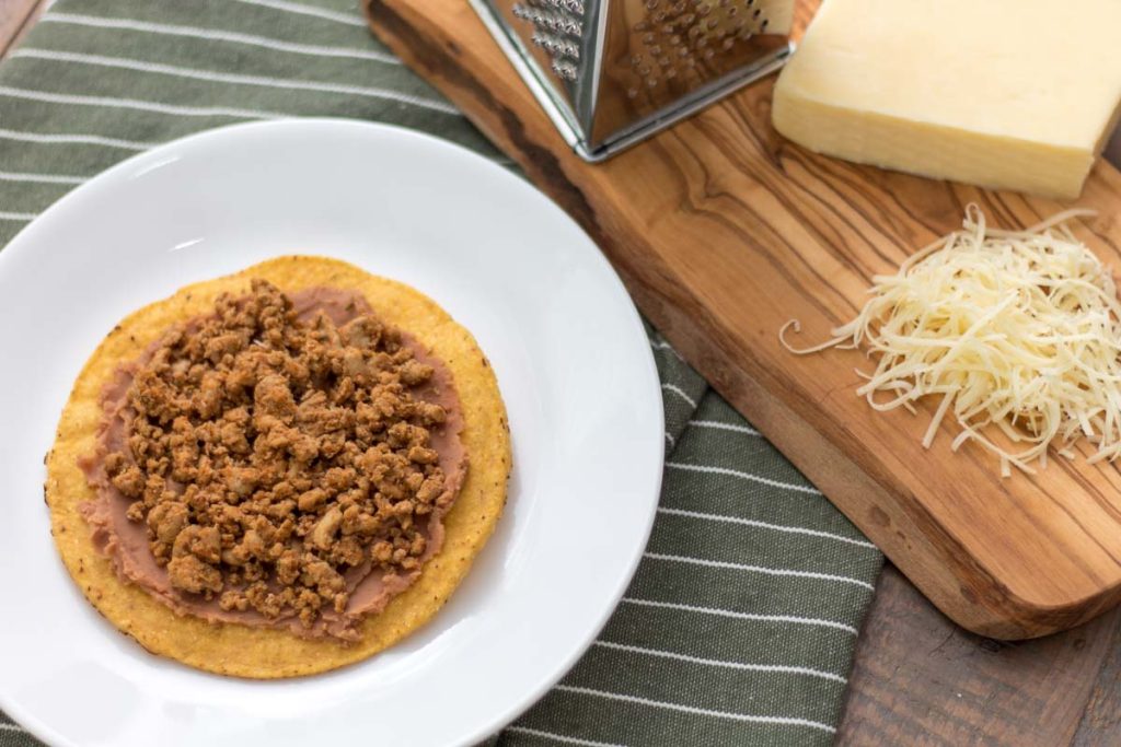 tostada shell with refried beans and taco meat on a plate next to freshly grated raw cheddar cheese and cheese grater on an olive wood cutting board