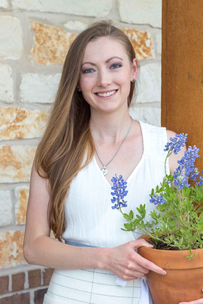 woman in white dress holding bluebonnets in a terracotta pot in front of a cedar column and limestone wall