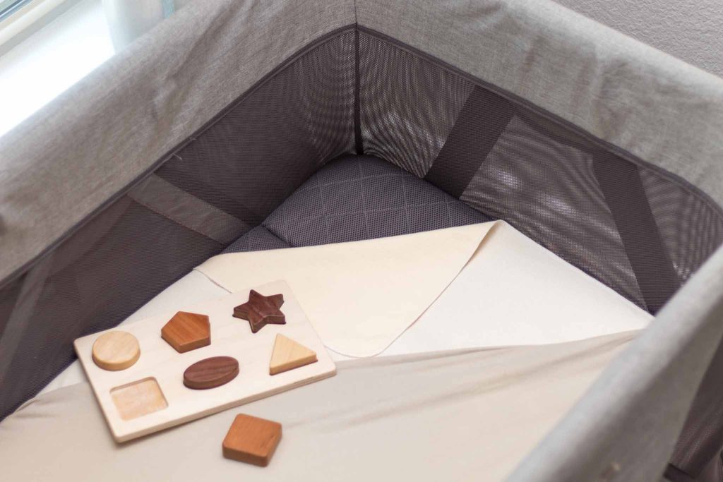 baby playard with mattress protector and sheet turned down in one corner with a natural wood shapes puzzle sitting on top