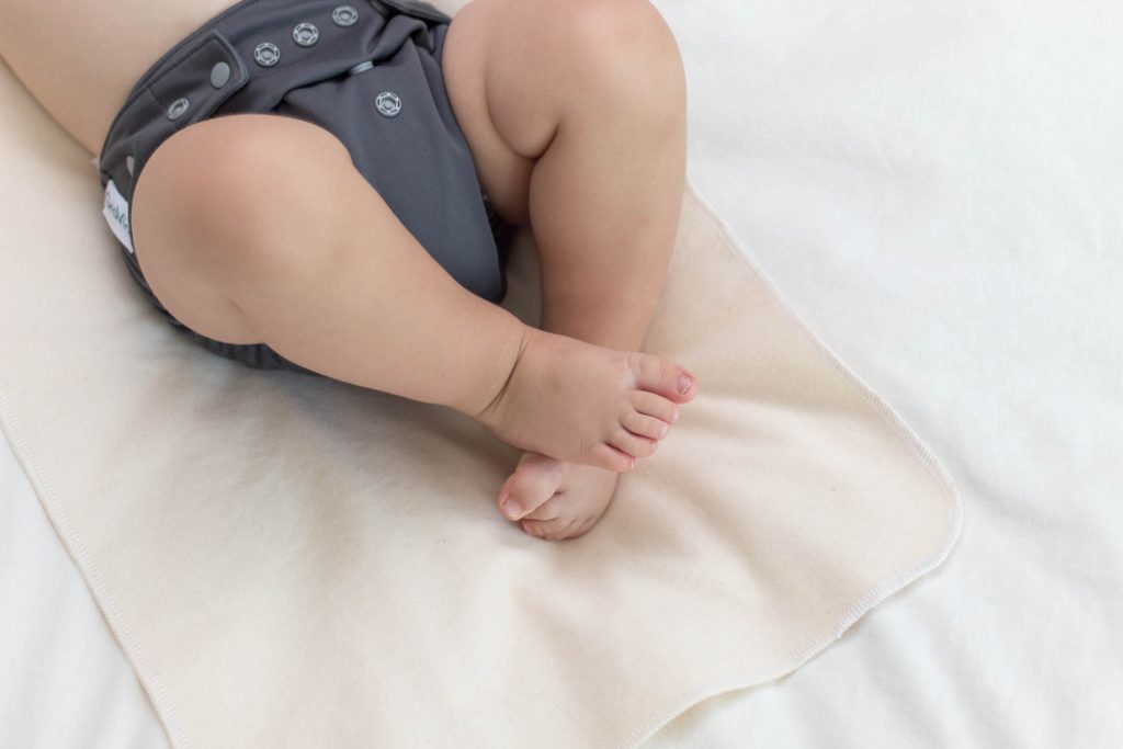 baby laying on changing mat (bassinet waterproof mattress protector) in a gray cloth diaper with legs curled up on top of an ivory down comforter on bed