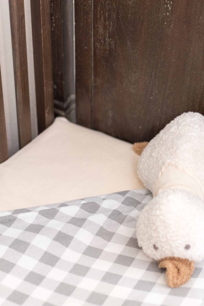 antiqued brown crib with gray and white buffalo check crib sheet turned down in one corner to reveal waterproof mattress protector with organic stuffed animal duck on top