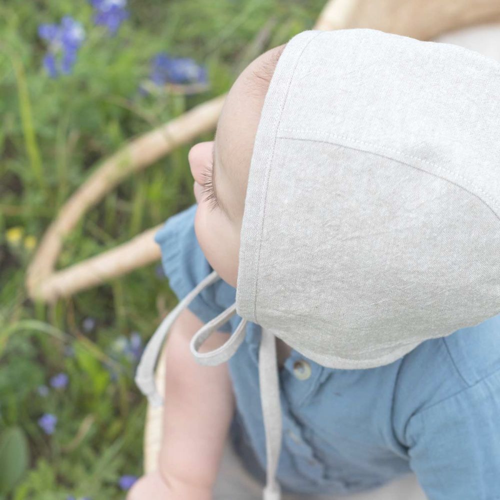 baby wearing a beige linen baby bonnet with muted blue gauze coverall in a moses basket bassinet in a field of Texas bluebonnets