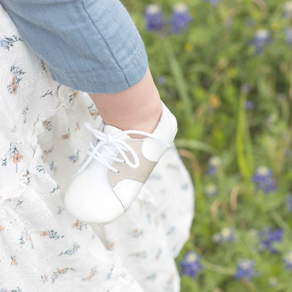 mother in white dress with small florals holding a baby wearing white and beige saddle shoes with muted blue gauze coverall in a field of Texas bluebonnets