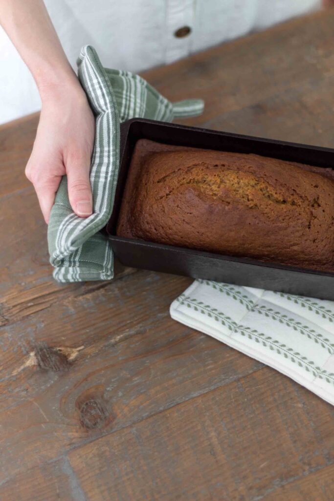 einkorn pumpkin bread in a cast iron loaf pan being placed on a farmhouse table with handmade pot holders