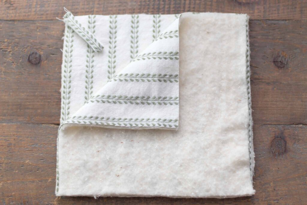 stacked squares of fabric with right sides facing together and a square piece of cotton quilt batting on top. left top corner folded down to show the fabric layers and folded loop placed in corner