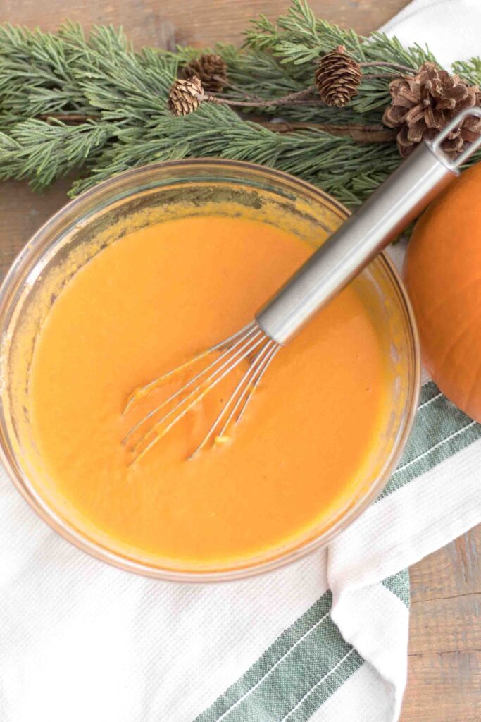 wet ingredients - eggs, oil, water, and pumpkin puree - mixed together in a glass bowl pictured with a whisk, pumpkin, and garland