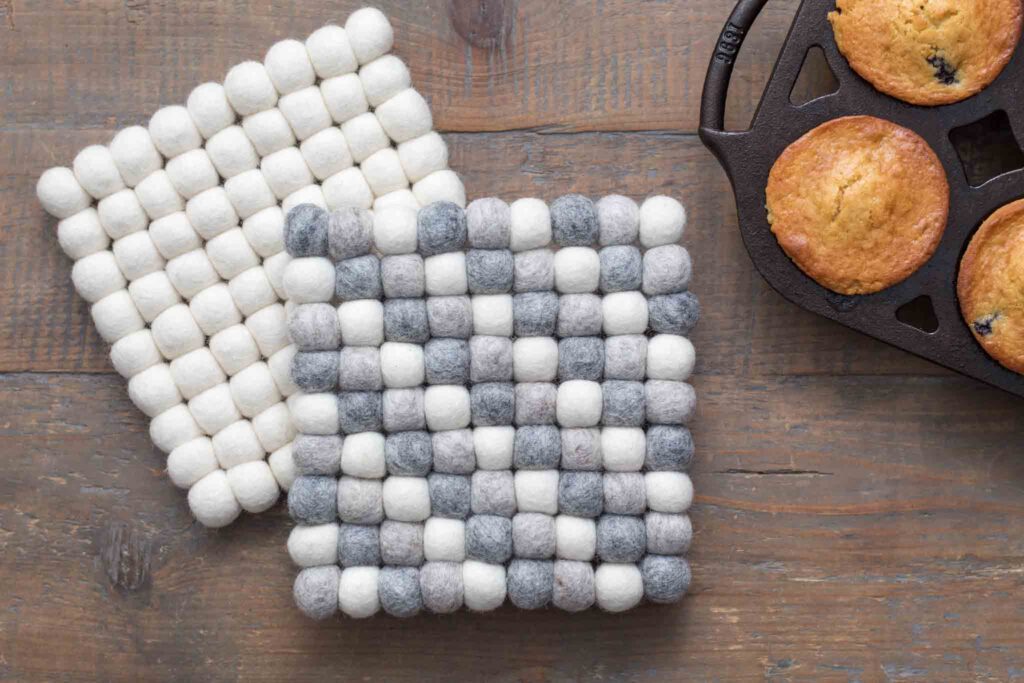close-up of two square wool felt ball trivets on a farmhouse table with homemade blueberry muffins in a cast iron loaf pan