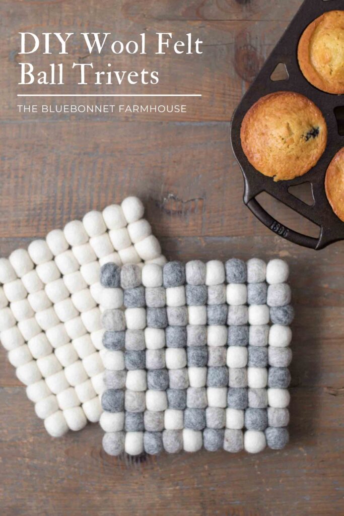two square wool felt ball trivets on a farmhouse table with homemade blueberry muffins in a cast iron muffin pan. colors of the trivets are ivory and natural gray wool. text reads "diy wool felt ball trivets"