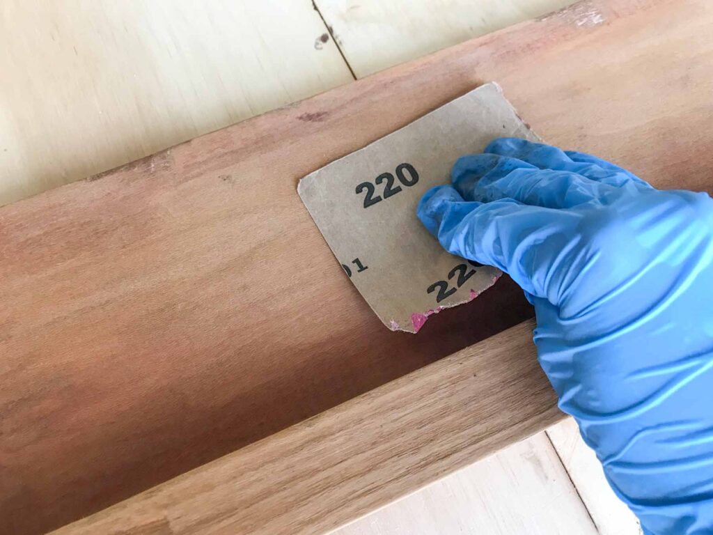 close-up photo of hand sanding antique bed side rails with 220 fine grit sandpaper after the wood conditioner has dried to remove any raised grains