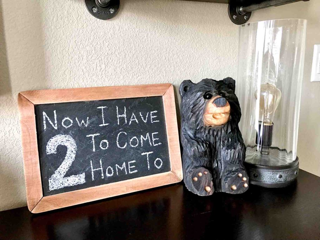 chainsaw carved black bear sitting on top of a dresser with an Edison light bulb lamp and small slate chalkboard that reads "now I have 2 to come home to".