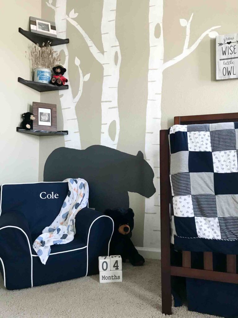 corner view of the woodland nursery reveal with a plush reading chair, DIY floating corner shelves, and white birch trees and black bear painted on the wall
