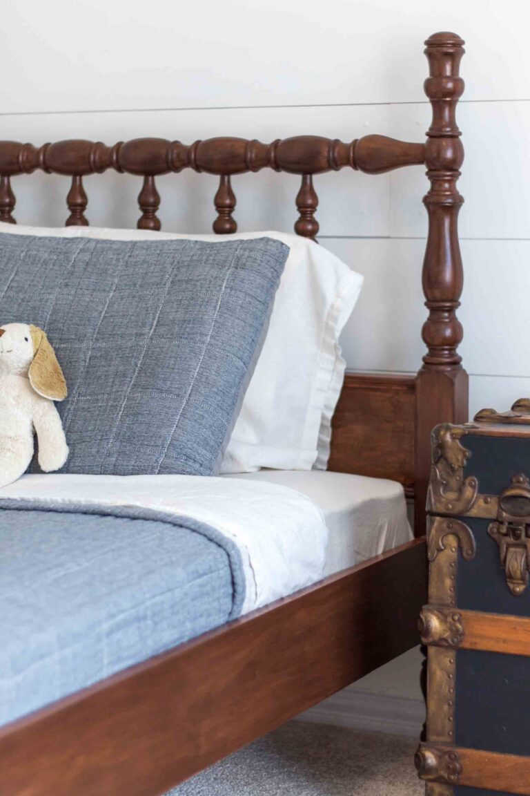 an antique jenny lind bed in front of a white shiplap wall with dusty blue and ivory bedding and an antique black steamer trunk