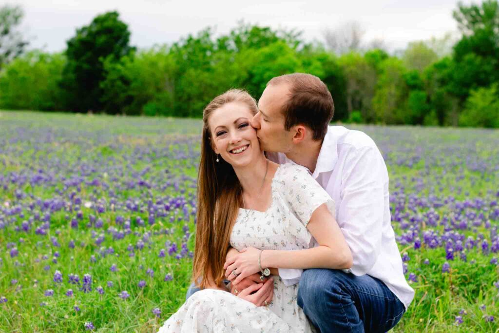 husband and wife dressed in white sitting in a field of bluebonnets. husbands arms wrapped around wife and kissing her on the cheek