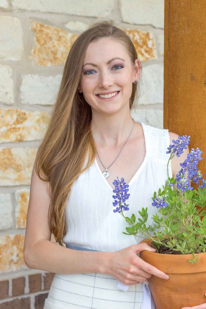 woman in white dress holding bluebonnets in a terracotta pot in front of a cedar column and limestone wall