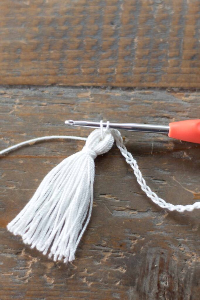 close-up of crochet hook pulling up a loop through the center of the tassel to attach with a single crochet to the chain tail of the cross bookmark