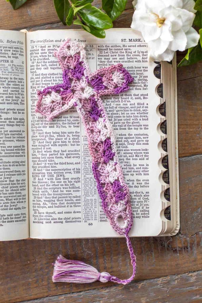flat lay of a lace crochet thread cross bookmark with tassel in shades of purple lying on top of an open bible in Mark chapter 15 next to a gardenia on top of a reclaimed farmhouse table