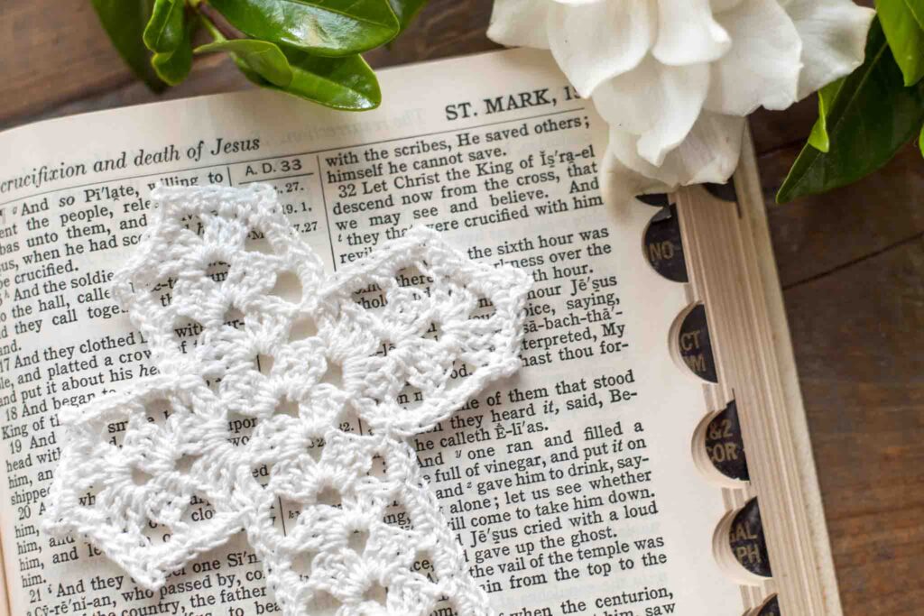 close-up of a white lace crochet thread cross bookmark on top of an open bible in Mark chapter 15 next to a gardenia on top of a reclaimed farmhouse table