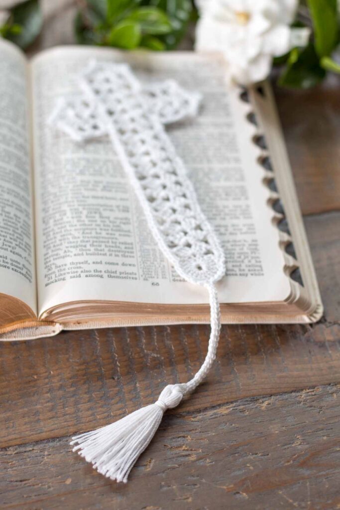 close-up of the tassel on a white lace crochet thread cross bookmark on top of an open bible in Mark chapter 15 next to a gardenia on top of a reclaimed farmhouse table