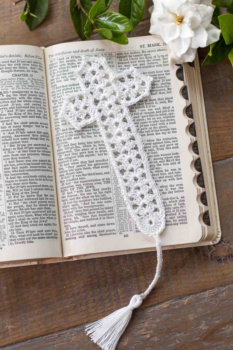 flat lay of a white lace crochet thread cross bookmark with tassel lying on top of an open bible in Mark chapter 15 next to a gardenia on top of a reclaimed farmhouse table