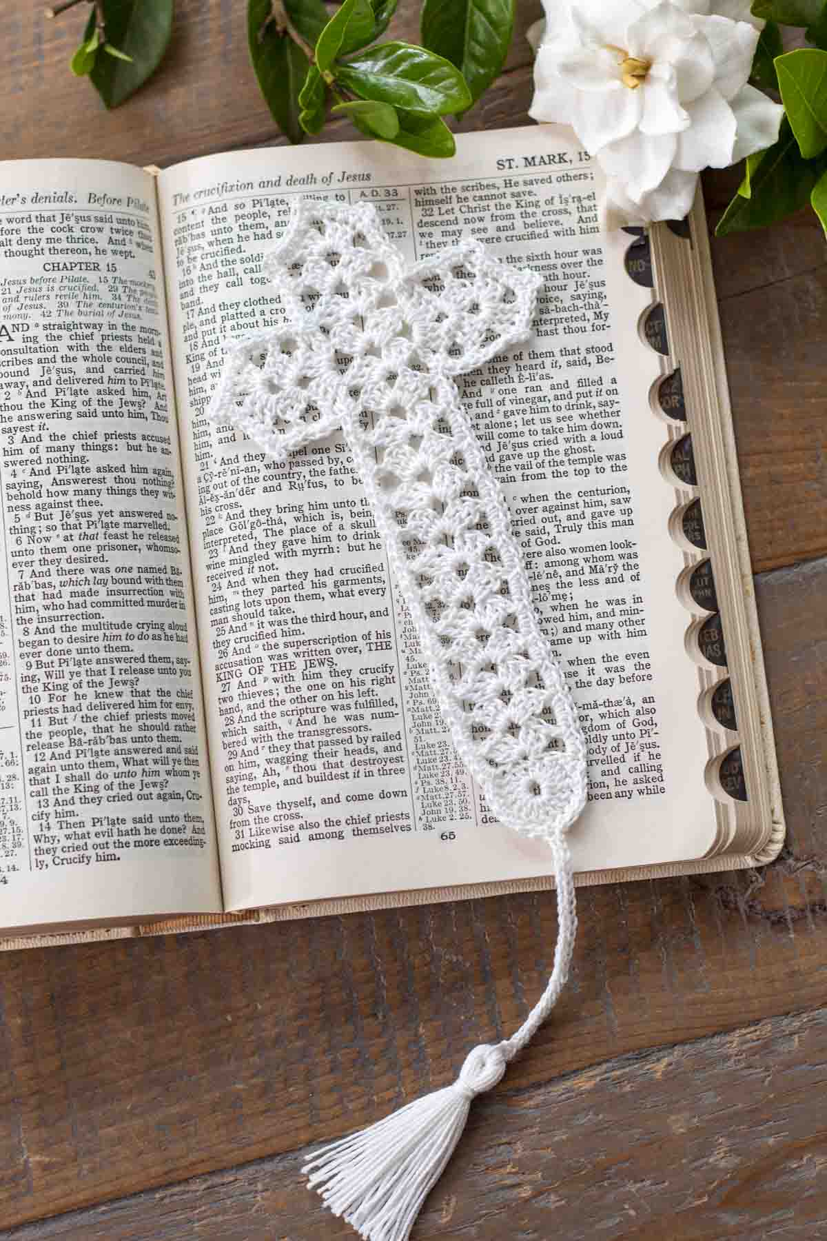 flat lay of a white lace crochet thread cross bookmark with tassel lying on top of an open bible in Mark chapter 15 next to a gardenia on top of a reclaimed farmhouse table