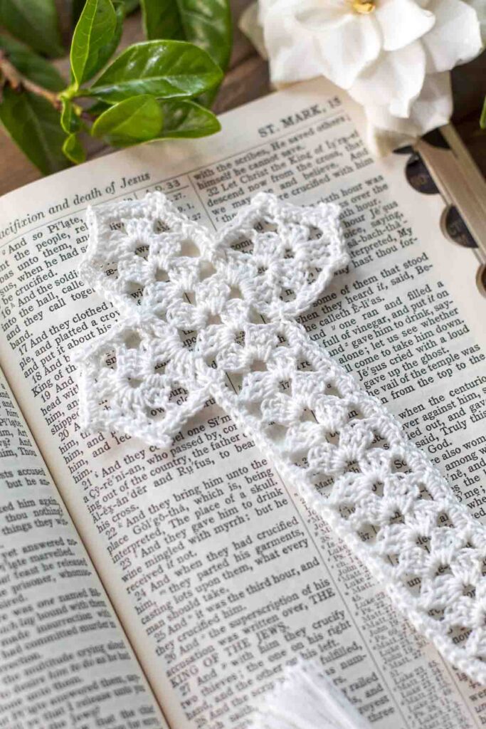 close-up of a white lace crochet thread cross bookmark with tassel lying on top of an open bible in Mark chapter 15 next to a gardenia on top of a reclaimed farmhouse table