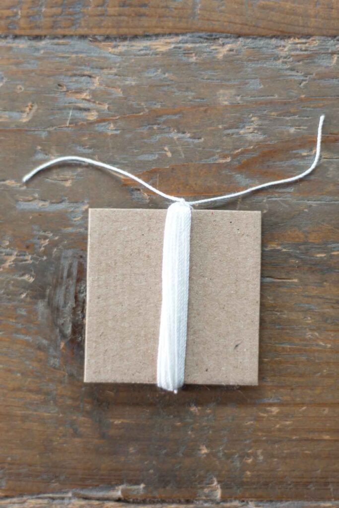 white crochet thread wrapped around a 2 inch by 2 inch square of cardboard 25 times with a knotted strand at the top of the tassel
