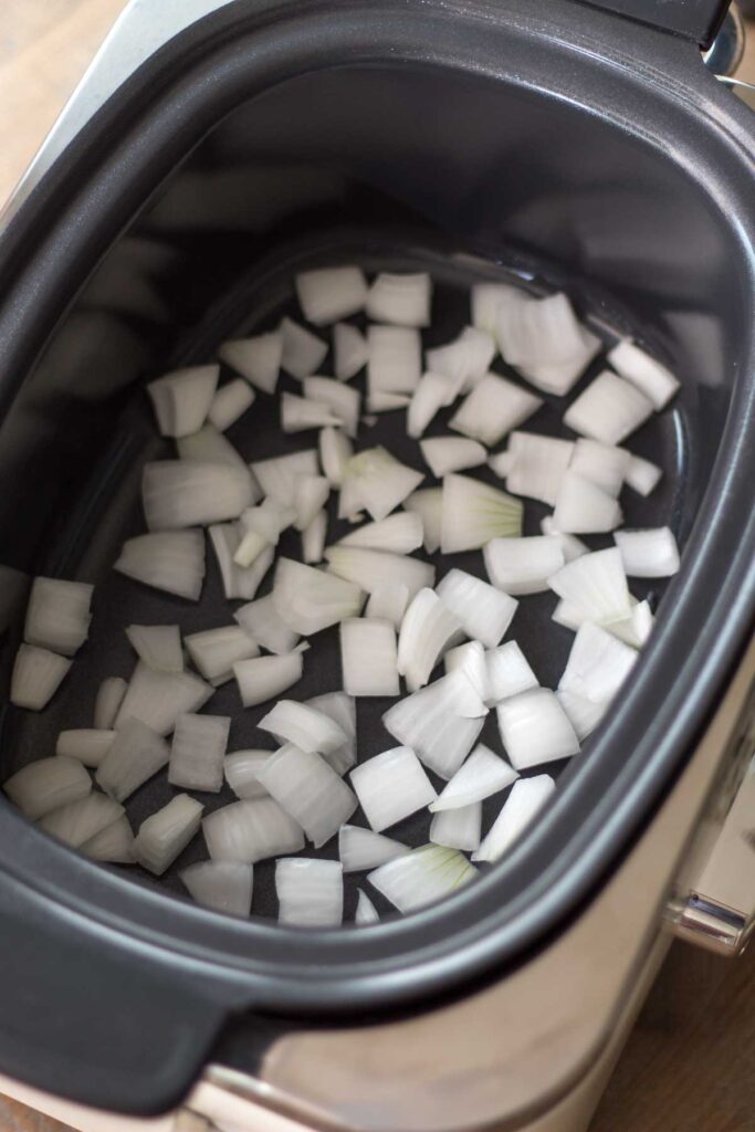 roughly chopped white onion scattered across the bottom of a slow cooker insert
