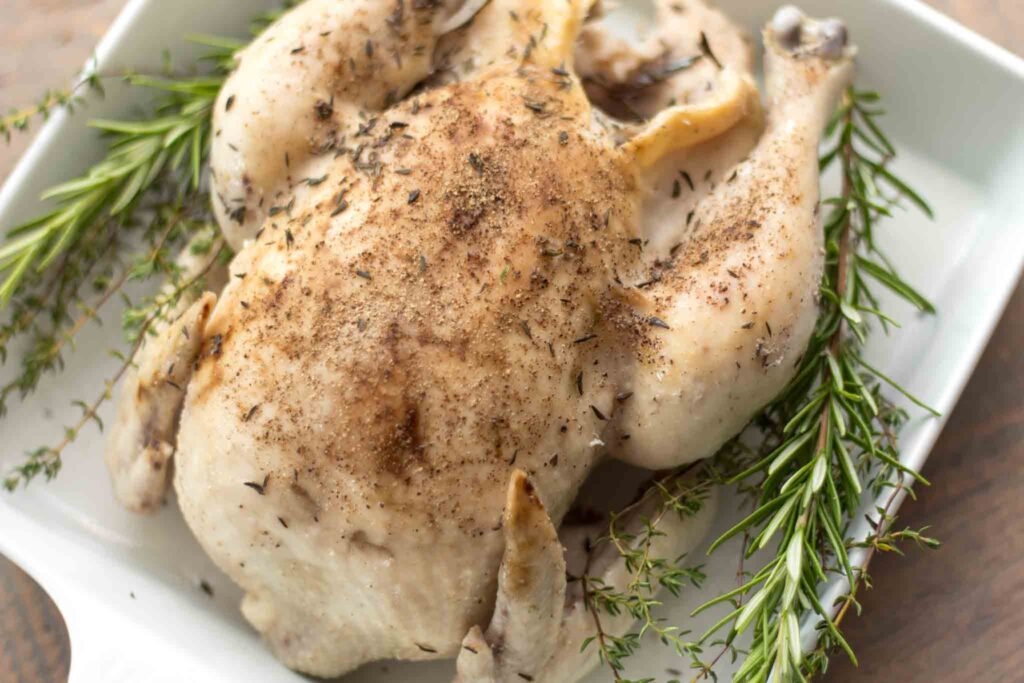 slow cooked whole chicken covered in salt and pepper with fresh sprigs of rosemary and thyme on each side in a white baking dish