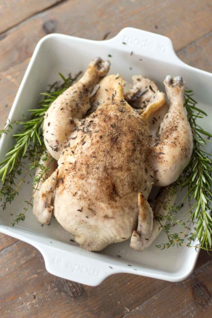 slow cooker whole chicken covered in salt and pepper with fresh sprigs of rosemary and thyme on each side in a white baking dish on a farmhouse table