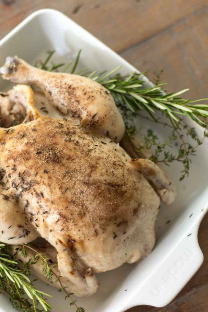 rosemary and thyme slow cooker whole chicken covered in salt and pepper with fresh sprigs of rosemary and thyme on each side in a white baking dish on a farmhouse table