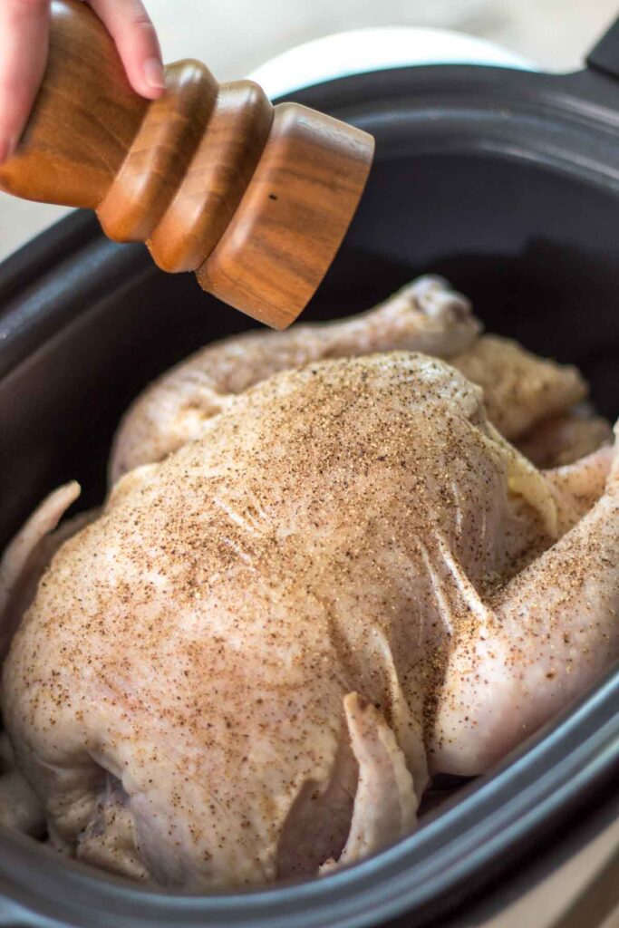 a walnut wood pepper mill sprinkling freshly ground black pepper across a whole chicken inside of a slow cooker