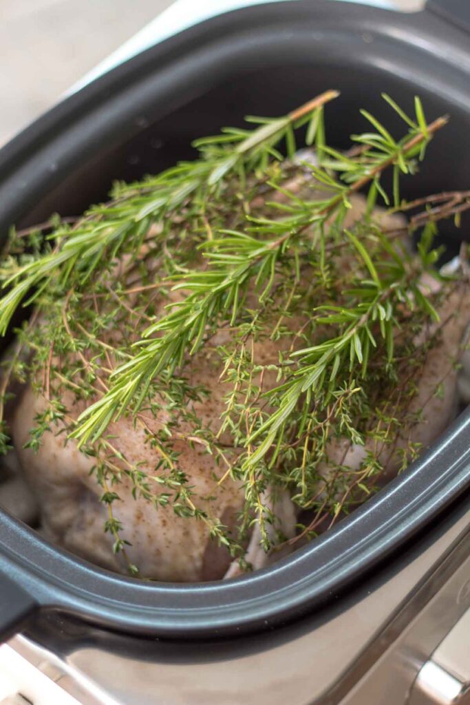 a handful of fresh thyme and rosemary sprigs lying across the top of a whole chicken in a slow cooker