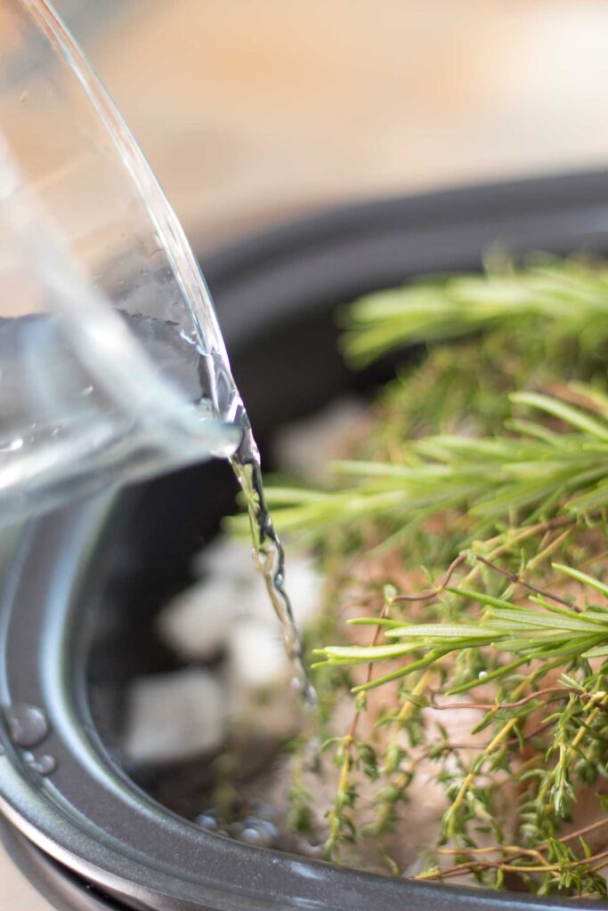 closeup of a stream of water pouring out from a glass measuring cup into a slow cooker filled with a whole chicken, onion, fresh rosemary and thyme
