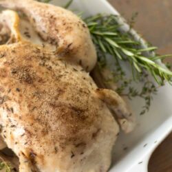 closeup of slow cooker whole chicken covered in salt and pepper lying on a bed of fresh rosemary and thyme in a white baking dish