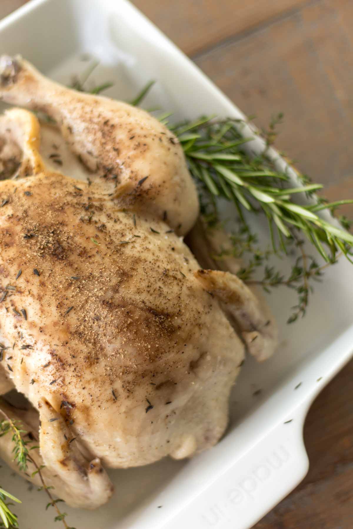 closeup of slow cooker whole chicken covered in salt and pepper lying on a bed of fresh rosemary and thyme in a white baking dish