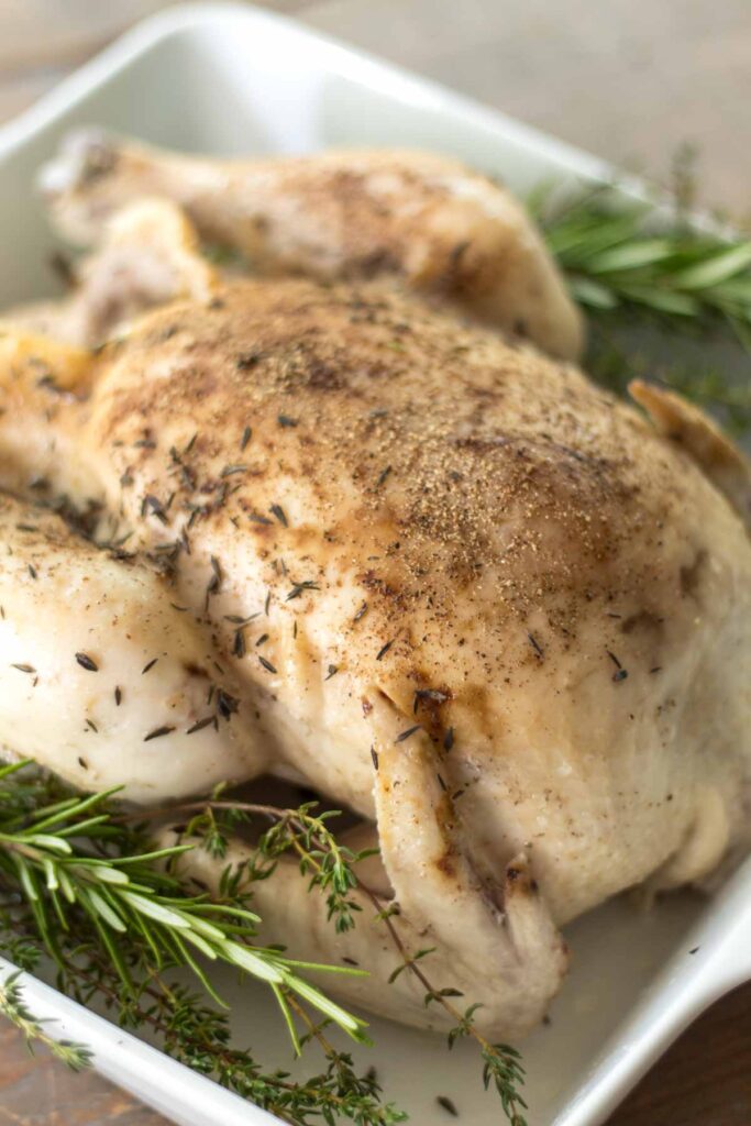 closeup of rosemary and thyme slow cooker whole chicken covered in salt and pepper with fresh sprigs of rosemary and thyme on each side in a white baking dish