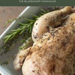 closeup of slow cooker whole chicken covered in salt and pepper lying on a bed of fresh rosemary and thyme in a white baking dish. text reads "rosemary and thyme slow cooker whole chicken -the bluebonnet farmhouse"