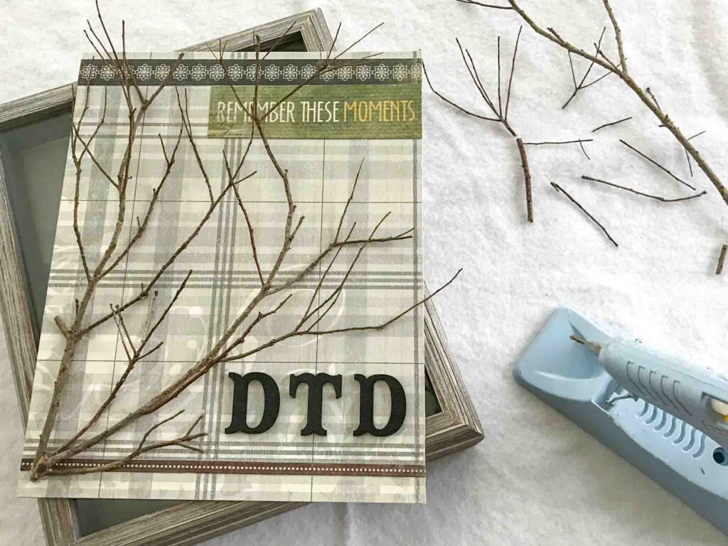 natural twigs on top of decorative neutral plaid scrapbook paper and floral trim with painted black wood letters for initials "DTD" on the back panel of a shadow box. decorative text reads "remember these moments" with a detail tip hot glue gun in the corner of the photo