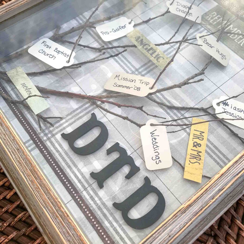 close up of a tree branch shadow box with painted black wood initials "DTD" and paper tags in the twigs with memories written on them.