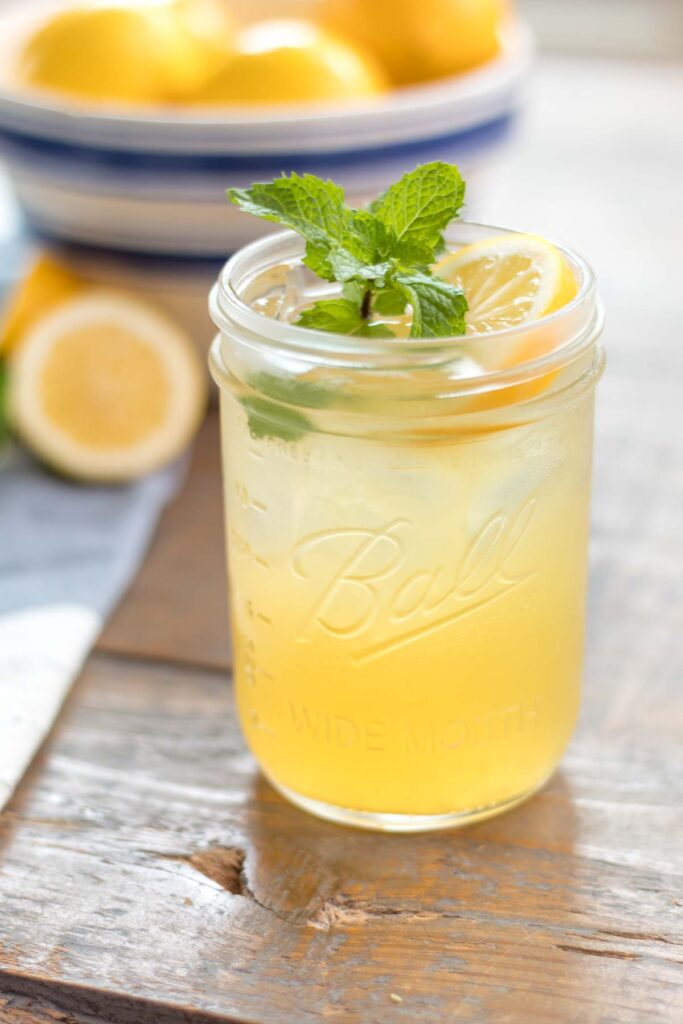 closeup of a glass wide mouth mason jar with iced homemade lemonade garnished with a fresh mint sprig and lemon slice. white with blue stripe antique stoneware bowl filled with lemons in the background