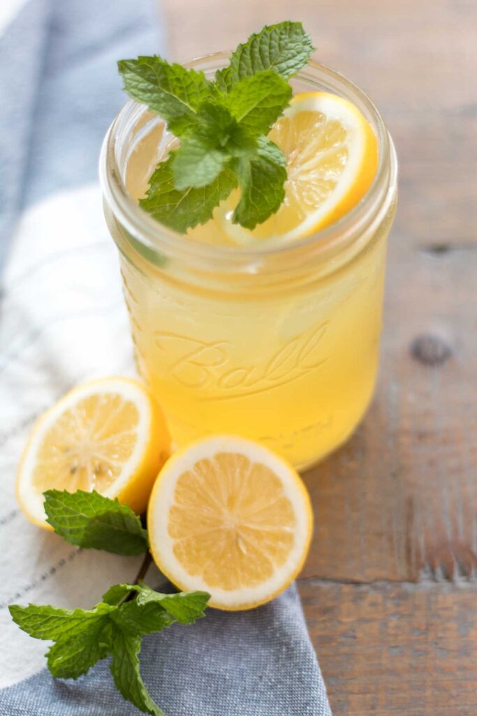 closeup of a glass wide mouth mason jar with iced honey lemonade garnished with a fresh mint sprig and lemon slice sitting next to a lemon cut in half and mint sprig on a blue and white flower sack towel